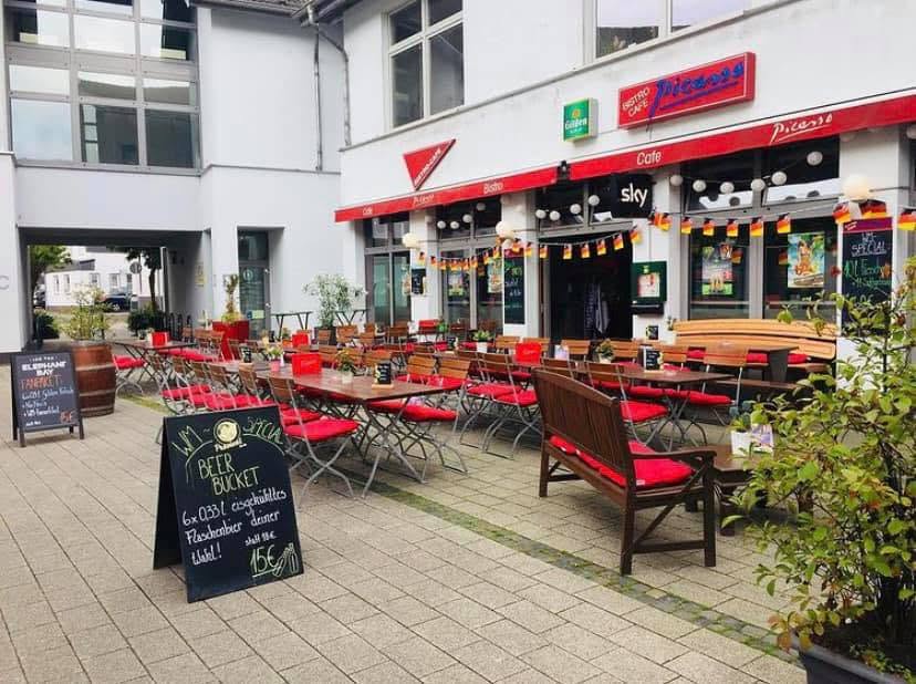 Terrasse vor dem Café Bistro Picasso mit roten Stühlen und Außentischen.